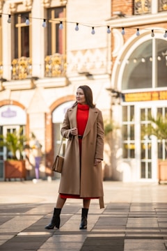 A person stands confidently in an urban setting, wearing a beige long coat over a bright red dress. They are accessorized with a tan handbag and black boots. The background features an elegant building with arched windows and decorative elements, while string lights hang above.