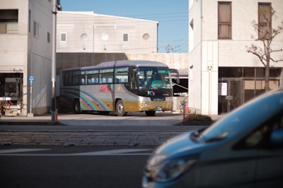 A large passenger bus with colorful stripes is parked near a modern building. The scene includes a partially visible car in the foreground and urban infrastructure such as a street and sidewalks.