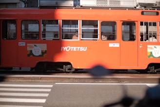 A vibrant orange tram with the text 'IYOTETSU' on its side is stationary at a tram stop. Passengers are visible through the windows, seated inside. The tram is partially in shadow with sunlight illuminating part of it, and there are advertisements featuring fruit on the side. In front of the tram, a zebra crossing can be seen.