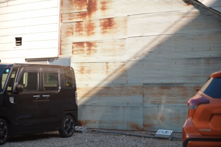 Two cars are parked in front of a corrugated metal wall, which shows signs of rust. The ground is covered with gravel, and there is a sign placed on the ground next to the cars.