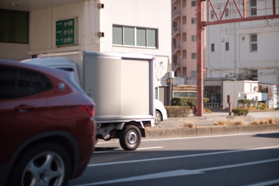 A small white truck is parked on a city street with an orange or brown car passing by in the foreground. There are buildings in the background along with street signs and some sparse vegetation.