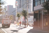 A city street scene with a cyclist in the foreground and a truck carrying gas containers on the left side of the road. There are several buildings, including a store with a 7-Eleven sign, and a few trees lining the street. The lighting suggests a sunny day, and some cars are parked along the side.