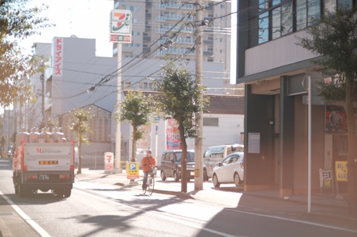 A city street scene with a cyclist in the foreground and a truck carrying gas containers on the left side of the road. There are several buildings, including a store with a 7-Eleven sign, and a few trees lining the street. The lighting suggests a sunny day, and some cars are parked along the side.