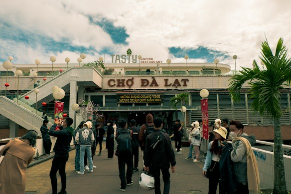 A bustling scene in front of a marketplace with a large sign reading 'Chợ Đ&agrave; Lạt.' Numerous people, some taking photos and others walking, are present. Lanterns and festive decorations are visible near the entrance. A restaurant sign on the upper level advertises Tasty Coffee Restaurant, and there is a clear sky in the background.