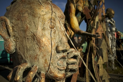 A collection of abstract sculptures made of metal and other materials, resembling human figures and faces. The weathered, textured surface of the large head in the foreground suggests uniqueness and creativity.
