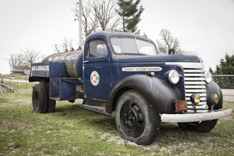 A vintage blue fuel truck with a rusted exterior is parked on a grassy area. The truck has an old-fashioned design with rounded headlights and a large cylindrical tank on the back. It bears logos and text indicating it's for gasoline delivery. Leafless trees and a house are visible in the background, contributing to a rustic setting.