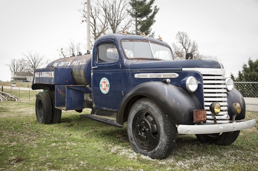 A vintage blue fuel truck with a rusted exterior is parked on a grassy area. The truck has an old-fashioned design with rounded headlights and a large cylindrical tank on the back. It bears logos and text indicating it's for gasoline delivery. Leafless trees and a house are visible in the background, contributing to a rustic setting.