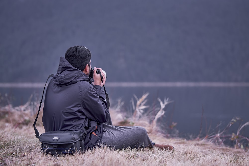 a man sitting in a field taking a picture of a lake