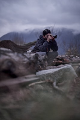 A person is crouched down, looking through the viewfinder of a camera, surrounded by natural elements such as logs and grass. The background features a slightly blurred mountain landscape with overcast skies.