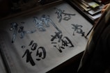 A person is practicing calligraphy on gridded paper, using a brush to write Chinese characters. The paper is placed on a table with assorted papers and possibly a book, partially visible in the background.