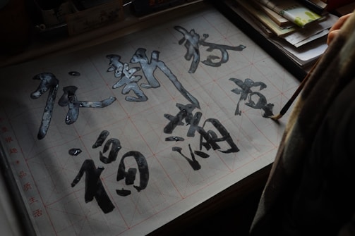 A person is practicing calligraphy on gridded paper, using a brush to write Chinese characters. The paper is placed on a table with assorted papers and possibly a book, partially visible in the background.
