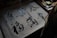 A person is practicing calligraphy on gridded paper, using a brush to write Chinese characters. The paper is placed on a table with assorted papers and possibly a book, partially visible in the background.