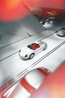 A sleek, silver vintage sports car is prominently displayed in a modern showroom with minimalistic architectural elements. The floor and walls are white, enhancing the sophisticated atmosphere. Red patches of light create a dynamic effect, contrasting with the neutral tones. Other cars are visible in the background, some partially obscured.