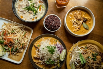 A variety of Thai dishes are arranged on a wooden table, featuring rich and colorful food. On the upper left, a bowl of creamy white soup is garnished with vegetables and oil droplets. Next to it, a bowl of reddish-brown rice is placed beside a beverage in a glass. The upper right features a bowl of curry with chunks of meat, spices, and a bay leaf. Below, a dish with a serving of Pad Thai has bean sprouts, a slice of lime, and crushed peanuts. Another curry dish is accompanied by rice noodles, herbs, and onions. To the left, a plate holds a vibrant papaya salad with tomatoes, beans, carrots, and peanuts.