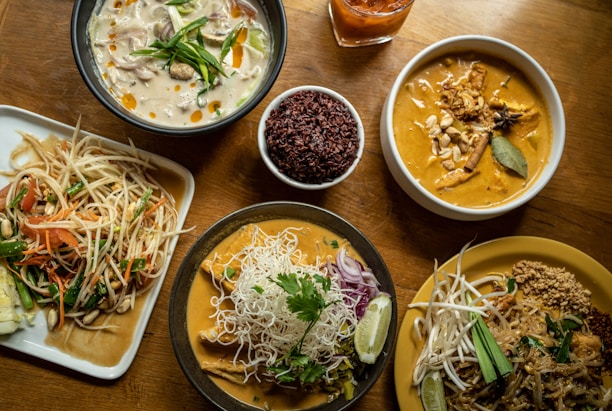 A variety of Thai dishes are arranged on a wooden table, featuring rich and colorful food. On the upper left, a bowl of creamy white soup is garnished with vegetables and oil droplets. Next to it, a bowl of reddish-brown rice is placed beside a beverage in a glass. The upper right features a bowl of curry with chunks of meat, spices, and a bay leaf. Below, a dish with a serving of Pad Thai has bean sprouts, a slice of lime, and crushed peanuts. Another curry dish is accompanied by rice noodles, herbs, and onions. To the left, a plate holds a vibrant papaya salad with tomatoes, beans, carrots, and peanuts.