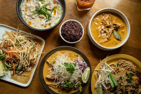 A variety of Thai dishes are arranged on a wooden table, featuring rich and colorful food. On the upper left, a bowl of creamy white soup is garnished with vegetables and oil droplets. Next to it, a bowl of reddish-brown rice is placed beside a beverage in a glass. The upper right features a bowl of curry with chunks of meat, spices, and a bay leaf. Below, a dish with a serving of Pad Thai has bean sprouts, a slice of lime, and crushed peanuts. Another curry dish is accompanied by rice noodles, herbs, and onions. To the left, a plate holds a vibrant papaya salad with tomatoes, beans, carrots, and peanuts.