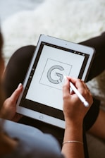 A person using a stylus to draw a geometric letter 'G' on a tablet, possibly designing a logo. The focus is on the screen, with the hand in the foreground holding the stylus.