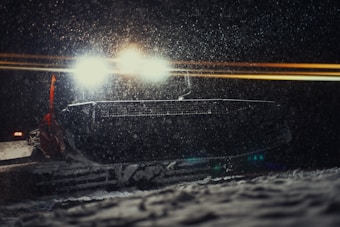 A snowplow operates at night, its bright headlights cutting through the falling snow. The vehicle's large front plow is visible amidst the snowfall, with a faint glow around it from the headlights. The scene suggests winter conditions with significant snowfall, creating a serene and isolated atmosphere.