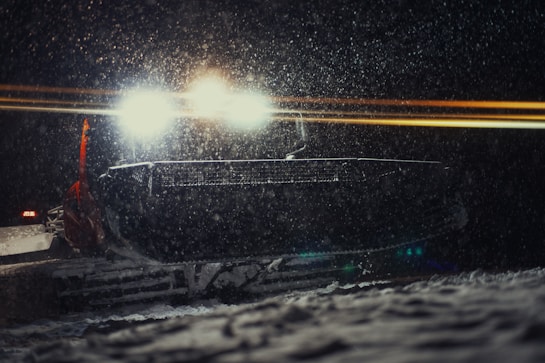 A snowplow operates at night, its bright headlights cutting through the falling snow. The vehicle's large front plow is visible amidst the snowfall, with a faint glow around it from the headlights. The scene suggests winter conditions with significant snowfall, creating a serene and isolated atmosphere.