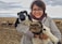 A smiling farmer standing beside a herd of sheep on a green pasture.