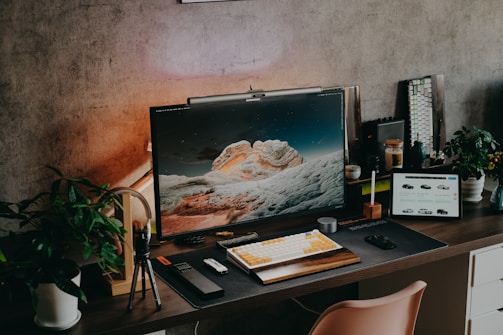 A modern desk setup featuring a large monitor displaying a scenic landscape with rocky formations under a starry sky. The workspace is neatly organized with a keyboard, mouse, a tablet showing a car selection website, and various decorative plants. There is ambient lighting creating a warm atmosphere, and additional tech gadgets and accessories are placed around the desk.