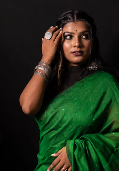 A graceful woman draped in a deep peacock green saree with golden embroidery, standing against a warm cream backdrop.