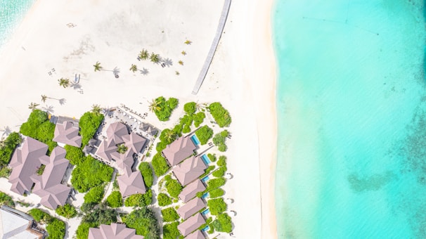 Aerial view of a beachside resort with several buildings surrounded by lush green vegetation. The sandy beach stretches parallel to the coast, leading to vibrant turquoise waters. Scattered palm trees provide a tropical atmosphere.