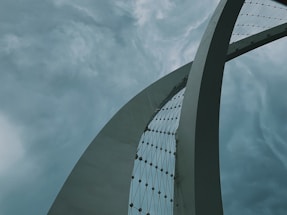 A large, sleek, and modern architectural structure curves gracefully against a dramatic sky. The structure features a network of cables, adding to its futuristic appearance. The sky is filled with heavy, swirling clouds, creating a dynamic and somewhat ominous backdrop.
