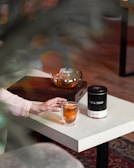 A person reaches for a glass mug of tea on a white table. Nearby, a black container labeled 'TEA SHOP' and a glass teapot sit on a wooden tray. The setting has a cozy and relaxed atmosphere.