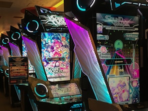 A row of brightly lit arcade machines with colorful screens and graphics. The machines feature the game Sound Voltex, with vibrant anime-style artwork and neon lights enhancing their appearance. The colors are bold and the machines are lined up side by side in a gaming arcade setting.