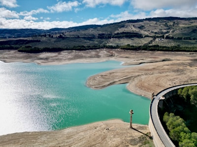 A reservoir with low water levels is surrounded by dry, rocky terrain and distant hills covered with patches of vegetation. The water appears to be a striking turquoise color, reflecting sunlight. A dam with a concrete structure is visible to the right, and sparse greenery is present near the base of the dam.