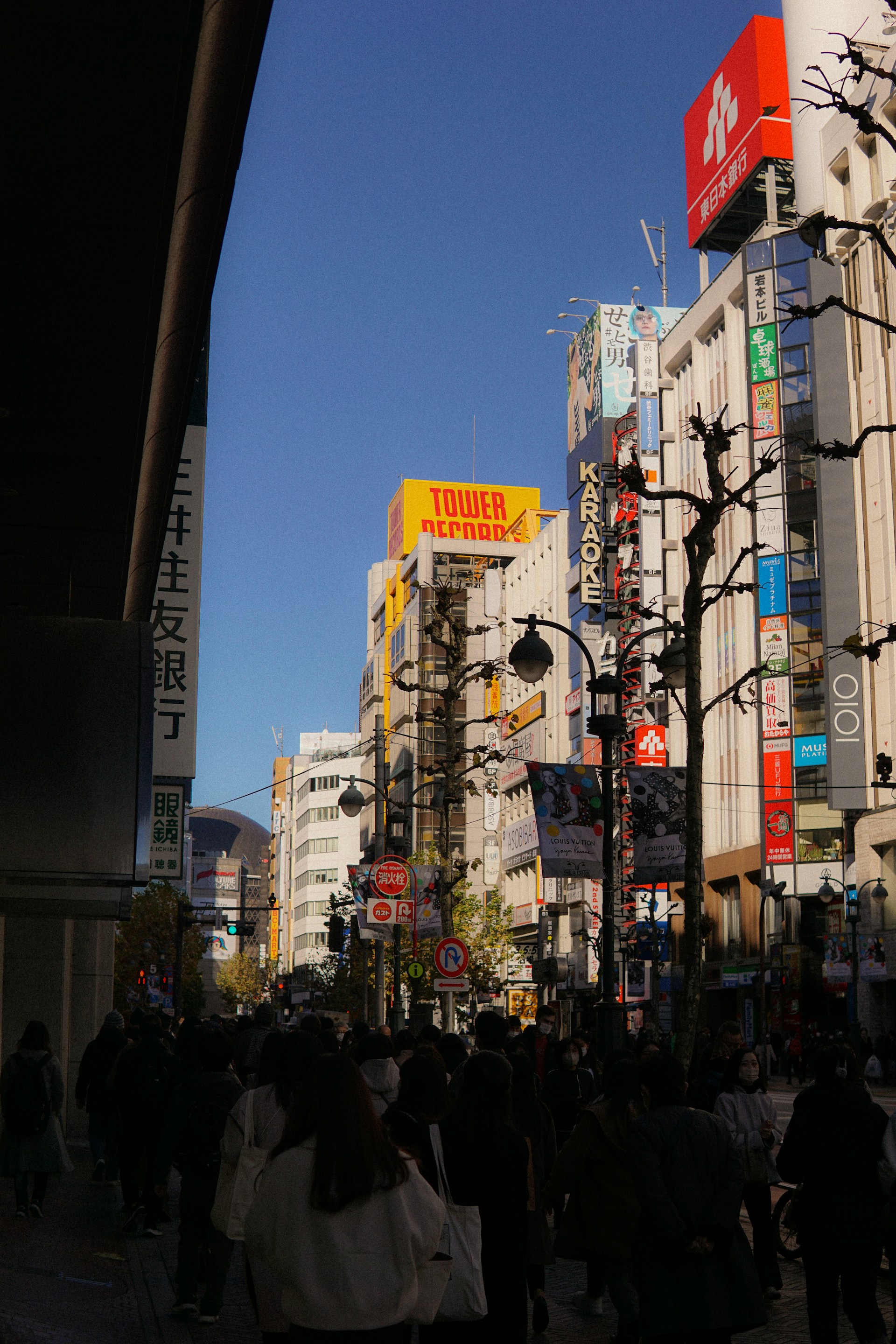 A bustling urban street scene with a crowd of people walking in the foreground. The background features high-rise buildings adorned with colorful billboards and signs, including a prominent yellow and red sign for a music store. Leafless trees line the street, and a clear blue sky provides a crisp backdrop.