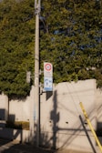A concrete utility pole stands by a sidewalk bordered with a low concrete wall and greenery. Two signs are attached to the pole: one indicates a speed limit of 30, and the other appears to be a pedestrian zone sign. Shadows from overhead wires and the pole itself stretch across the sidewalk.
