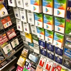 A display rack featuring a variety of gift and prepaid cards from popular brands. Cards are organized in stacks and rows, including Google Play, Apple iTunes, Amazon, PlayStation, Netflix, and more. The colors are vibrant and the packaging is clearly branded.