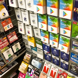 A display rack featuring a variety of gift and prepaid cards from popular brands. Cards are organized in stacks and rows, including Google Play, Apple iTunes, Amazon, PlayStation, Netflix, and more. The colors are vibrant and the packaging is clearly branded.