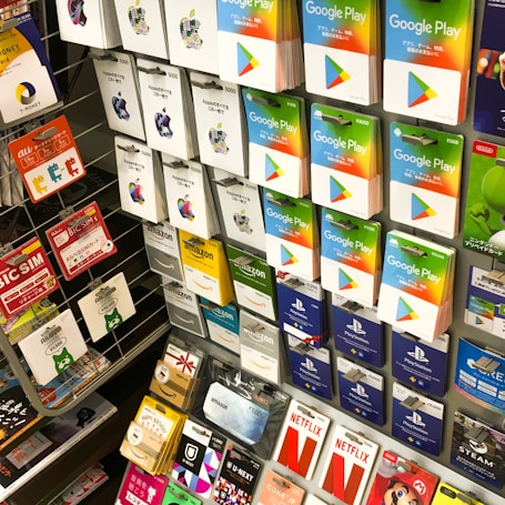 A display rack featuring a variety of gift and prepaid cards from popular brands. Cards are organized in stacks and rows, including Google Play, Apple iTunes, Amazon, PlayStation, Netflix, and more. The colors are vibrant and the packaging is clearly branded.