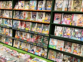 A bookstore shelf filled with various volumes of manga, displaying colorful covers featuring anime-style illustrations. The shelves are densely packed, and multiple rows showcase an array of genres. The titles and artwork reflect a vibrant and diverse selection typical in Japanese manga stores.