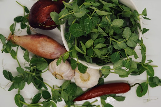 A collection of fresh vegetables and herbs is arranged on a white surface. There are pea shoots in a bowl, surrounded by a red onion, a garlic bulb, a shallot, and a red chili pepper.