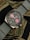 A wristwatch with a metal case and a gray fabric strap, featuring a dark dial with pink subdials and white hour markers. The watch has chronograph functions indicated by the subdials and text on the bezel. It is placed on a background with a camouflage pattern.