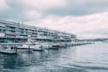 A waterfront scene with several luxury yachts docked at a marina. The background features a large, modern apartment building with multiple balconies and terraces. The sky is overcast with heavy clouds, casting a muted tone over the water.