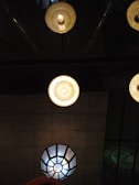 Several ceiling lights with different designs and brightness are visible. The foreground features two circular lamps with warm, glowing bulbs, while a geometric lamp with a cooler light is situated in the background. The ceiling is textured, adding depth to the overall scene.