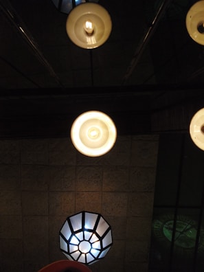 Several ceiling lights with different designs and brightness are visible. The foreground features two circular lamps with warm, glowing bulbs, while a geometric lamp with a cooler light is situated in the background. The ceiling is textured, adding depth to the overall scene.