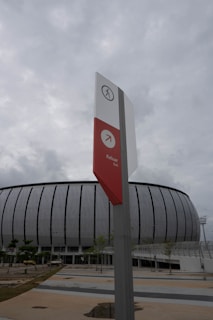 A modern, curved building with vertical lines on its facade is set against a cloudy sky. In the foreground, a tall directional sign stands upright, featuring a pedestrian icon at the top and an arrow pointing right labeled 'Keluar Exit' in red.