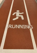 A section of a running track with a textured surface in a reddish-brown color. White lines border the lane, and a white pictogram of a person running is placed above the word RUNNING painted on the track.
