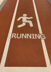 A section of a running track with a textured surface in a reddish-brown color. White lines border the lane, and a white pictogram of a person running is placed above the word RUNNING painted on the track.