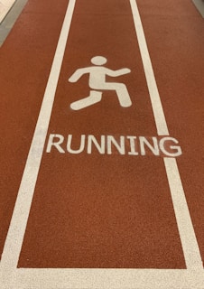 A section of a running track with a textured surface in a reddish-brown color. White lines border the lane, and a white pictogram of a person running is placed above the word RUNNING painted on the track.