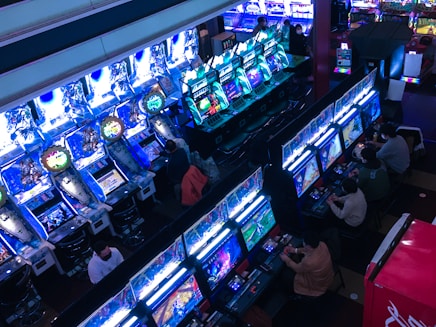 A bustling arcade filled with rows of gaming machines, each with vibrant displays and controls. Several people are seated at these machines, deeply engaged in playing games. Bright fluorescent lights cast a blue and neon glow across the room, enhancing the lively and energetic atmosphere.