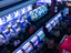 A bustling arcade filled with rows of gaming machines, each with vibrant displays and controls. Several people are seated at these machines, deeply engaged in playing games. Bright fluorescent lights cast a blue and neon glow across the room, enhancing the lively and energetic atmosphere.