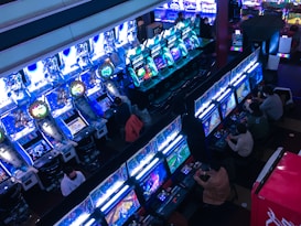 A bustling arcade filled with rows of gaming machines, each with vibrant displays and controls. Several people are seated at these machines, deeply engaged in playing games. Bright fluorescent lights cast a blue and neon glow across the room, enhancing the lively and energetic atmosphere.