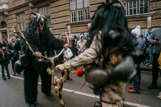 Two individuals wearing elaborate costumes resembling mythical creatures with fur, horns, and masks engage in an outdoor performance or festival. They carry wooden staffs decorated with small flowers. Spectators, dressed in winter clothing, stand around watching the event on a city street with historic brick buildings as the backdrop.